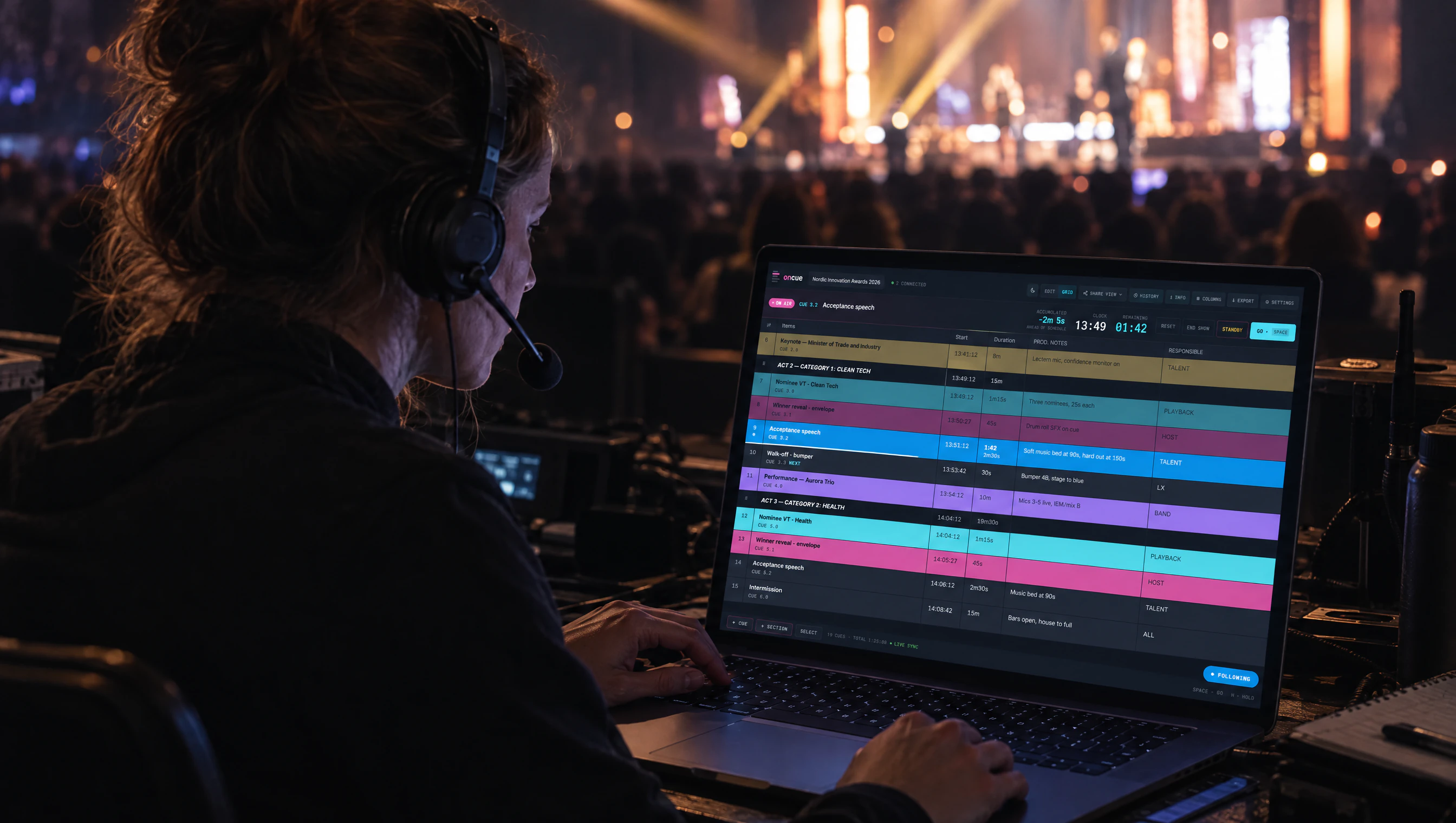 A show caller with a headset at the front-of-house desk, the OnCue editor open on a laptop and the lit stage and audience behind her