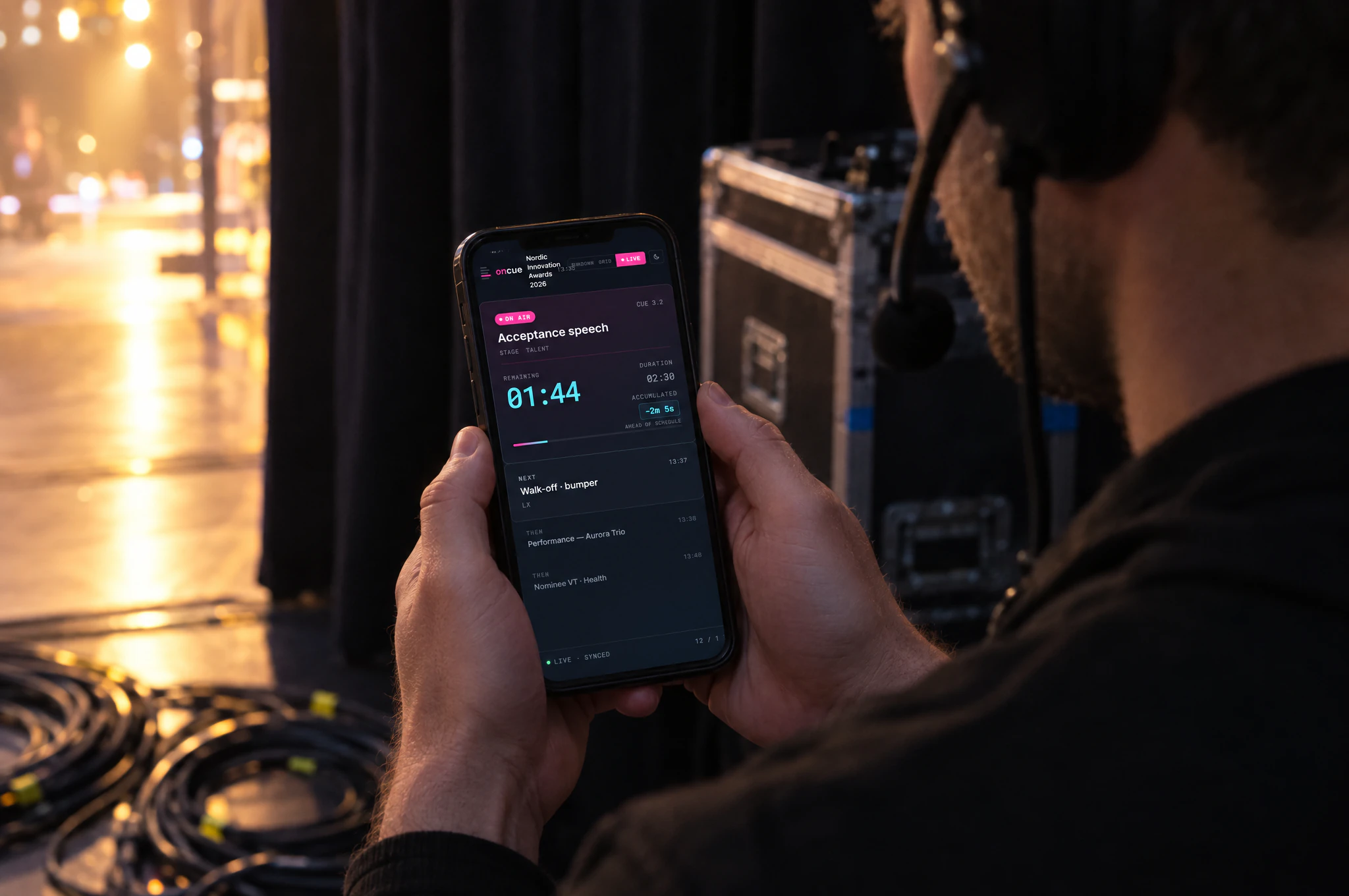 A stage manager in the wings holding a phone that shows the OnCue live viewer with the on-air cue and countdown